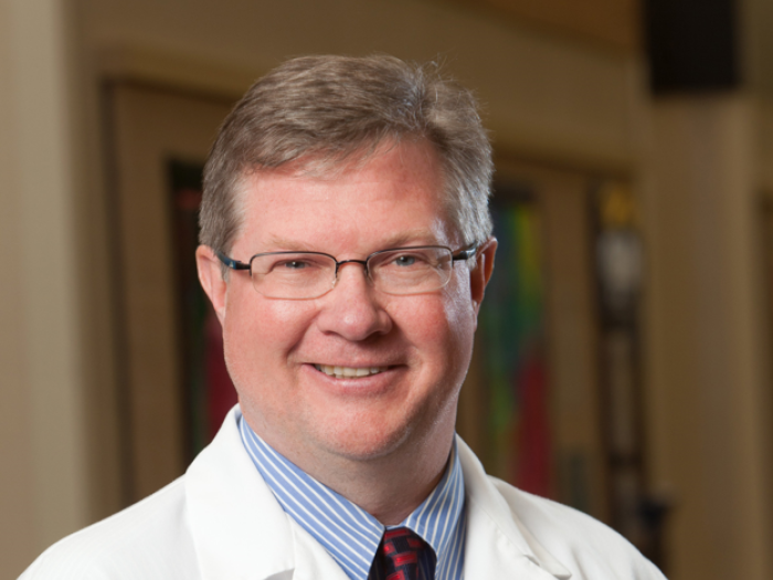 Professional portrait of a white middle aged man with short light brown hair, wearing glasses, a business shirt, a tie, and a white doctor's coat
