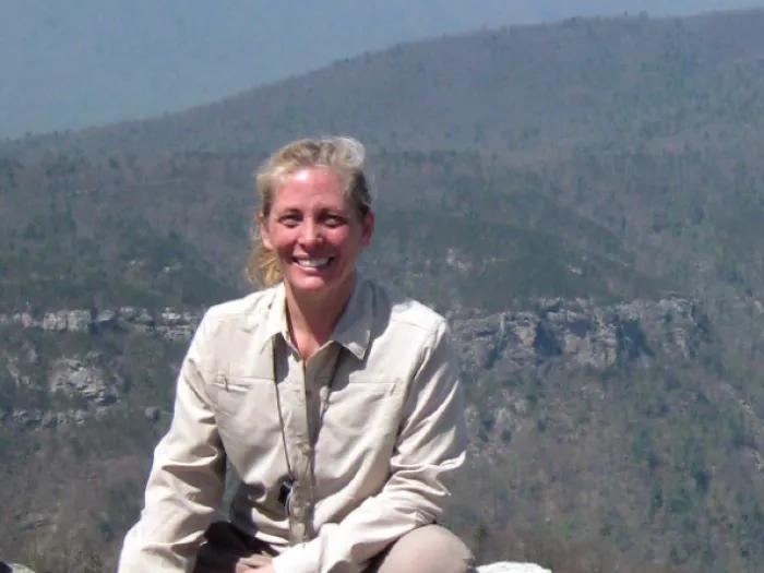 A white woman in khaki pants and shirt crouches on a rocky outcropping overlooking a mountain range