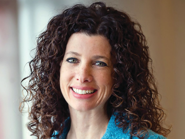 Professional portrait of a white woman with dark curly hair. She is wearing a blue blazer.
