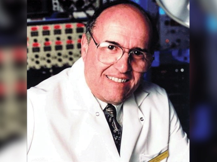 Portrait of an older man in a white lab coat wearing glasses and smiling. There's lab equipment in the background.