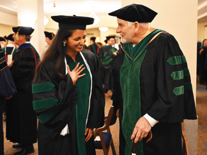 Two people in black and green graduation robes share a happy moment of celebration in a crowded lobby. The woman on the left is younger and smiling or laughing. The older man on the right is carrying a cane and smiling at the woman.