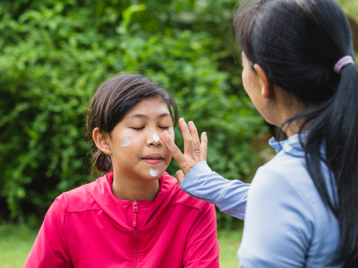 A mom applies sunscreen to her daughter's face. They are outdoors in a green area. Both are wearing long-sleeve jackets.