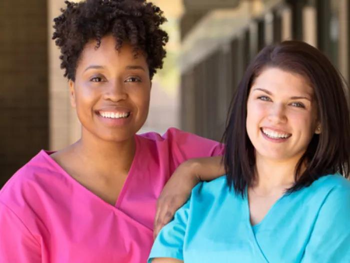 Stock image of two health care providers standing together outside. The one to the left is wearing pink scrubs. The one on the right is wearing light blue scrubs.