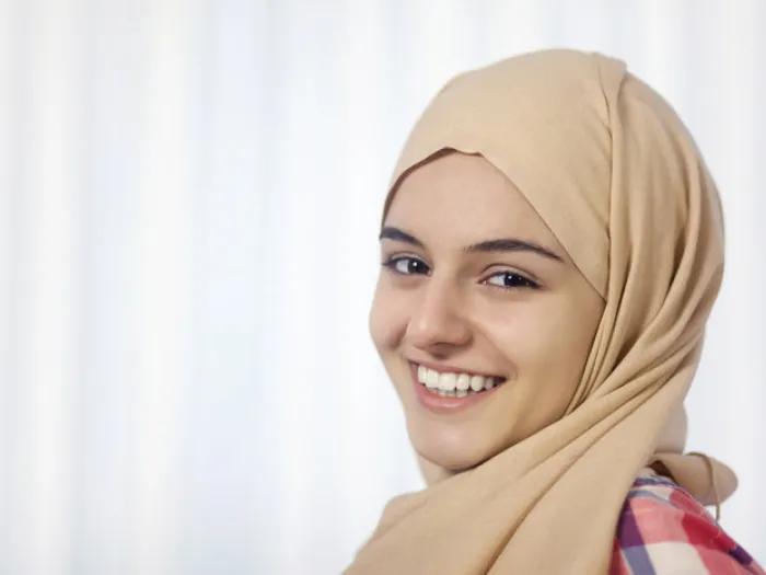 Stock image of a young person smiling. They are wearing a tan hijab and a plaid shirt.