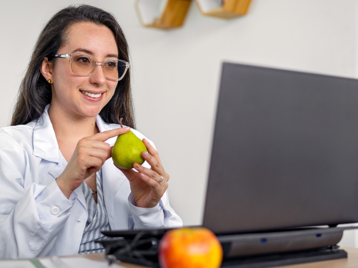 person in white coat at laptop green fruit in hand