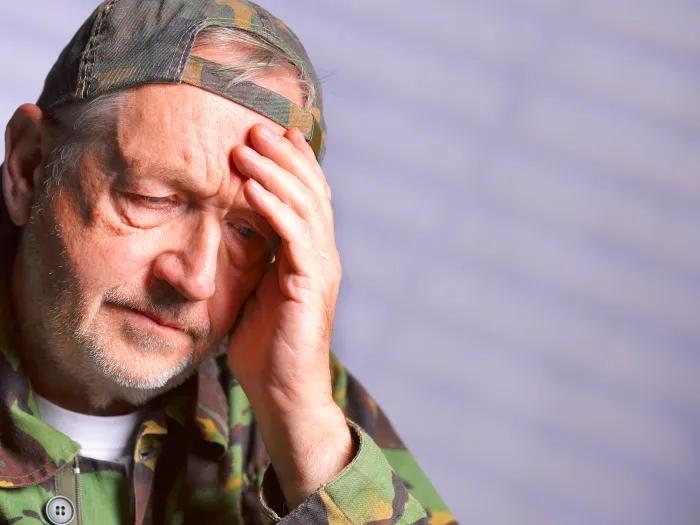 Older man with hand on forehead, dressed in camouflage clothing