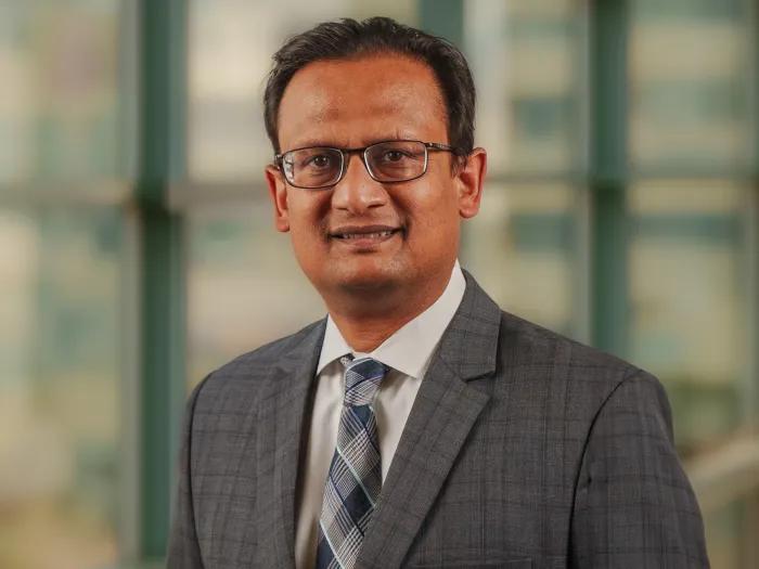 Dr. Nitin Jain standing in a hallway wearing a gray suit jacket and gray plaid tie