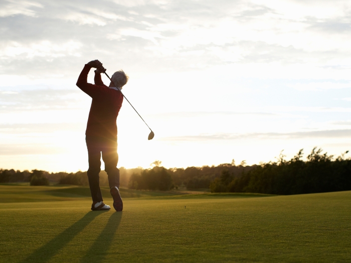 Older man swings a golf club with sunset in the background