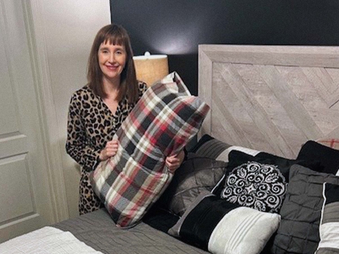 smiling woman holds pillow with plaid cover next to bed