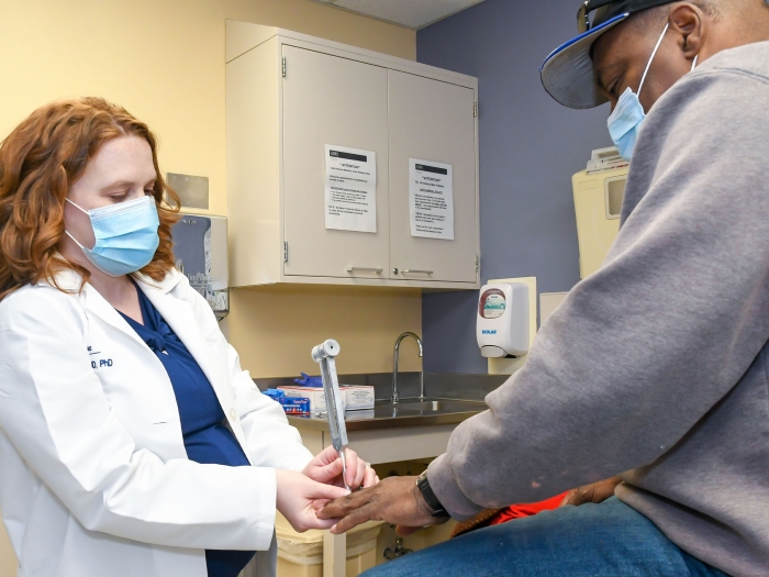 doctor in white coat with dark blue scrubs touching hand of patient in grey sweater and baseball cap in exam room 