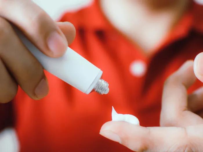 white cream being put on finger close up with blurred red shirt in background of person using cream