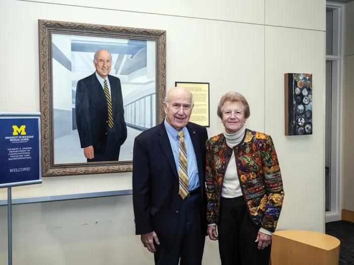 Gilbert Omenn and Martha Darling pose in front of a portrait of Omenn at the University of Michigan Medical School. Omenn is wearing a suit and tie and Darling is wearing a colorful print blazer.