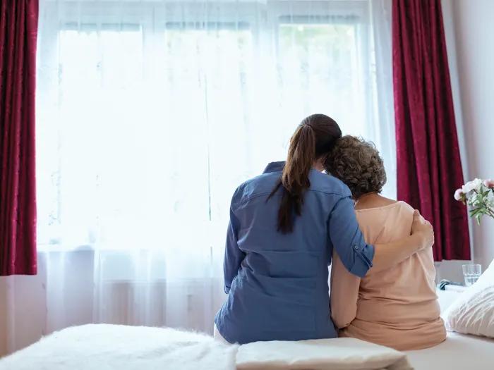 A hospice caregiver sits next to a patient on a bed. Both are facing away from the camera and look toward a window with diaphanous curtains that let in a lot of sunlight. The hospice worker has her arm around the patient, and the patient has her head on the hospice worker's shoulder.