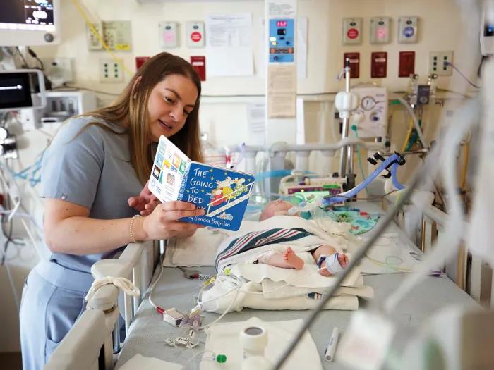 A nurse reads The Going to Bed Book by Sandra Boynton to a baby who is in the Neonatal Intensive Care Unit. The baby is in a hospital crib and hooked up to several machines. The nurse is wearing blue-gray scrubs.