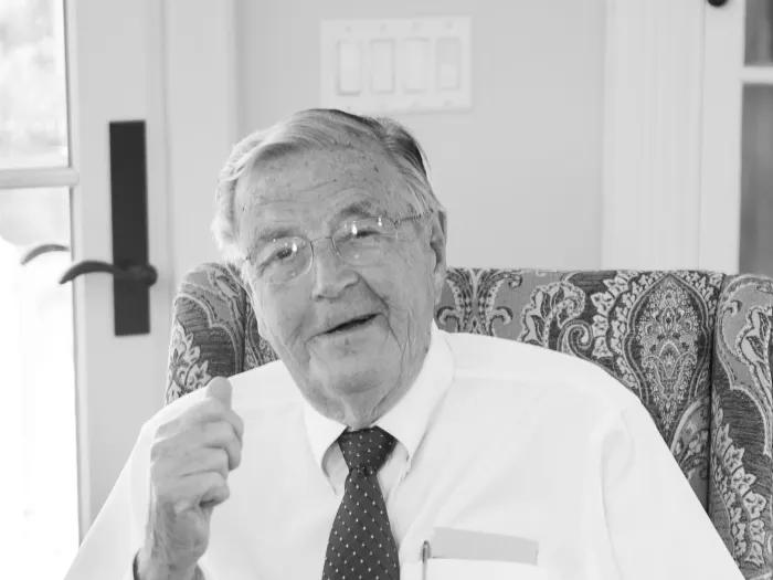Smiling black and white photo of Phil Jenkins seated at his desk. He is dressed up in a button-up shirt and tie.