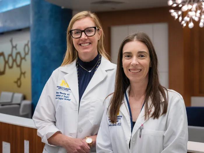 Melissa Pilewskie, M.D., associate professor of surgery, and Monika Burness, M.D., assistant professor of internal medicine, pose together on the Medical Campus. They are both wearing white coats, smiling, and looking at the camera.