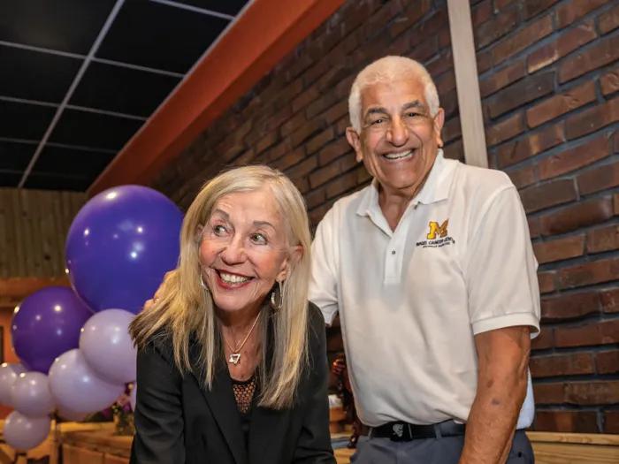 Susan and Richard Rogel are standing together behind a table. Susan is leaning over with a pen in her hand to sign a gift agreement. She's smiling and looking at someone out of the frame. Richard is smiling and looking directly at the camera.