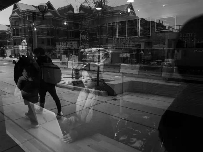 Black and white artistic photograph of a woman sitting inside a coffee shop working on a computer. The photo is taken from outside, so there's a reflection on the window of people walking down the street.
