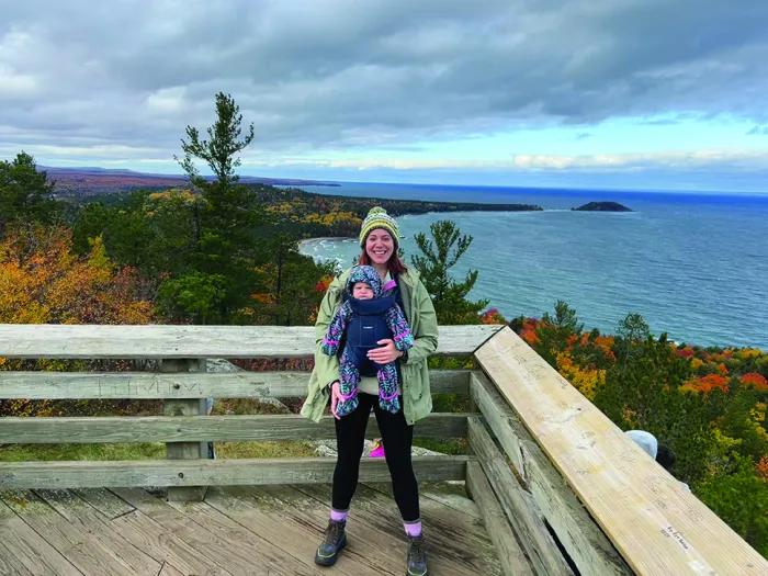 A young woman holding a baby poses in front of a beautiful vista in the Upper Peninsula. Visible in the background are the lake and trees just starting to change color in the fall.