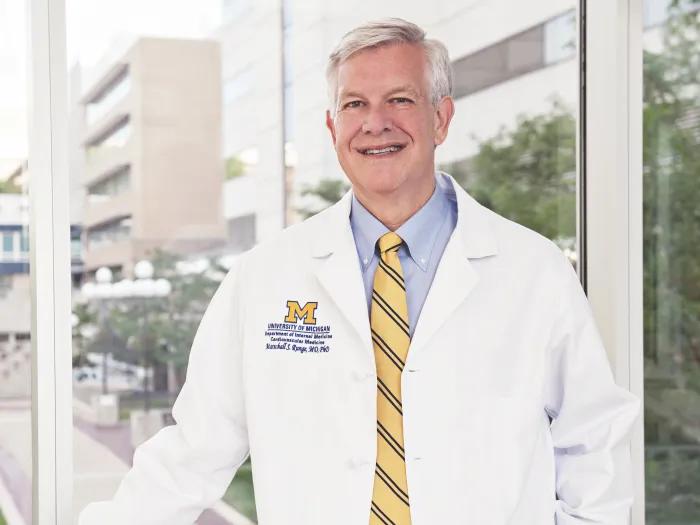 Marschall Runge is standing in front of a window wearing a doctor's whitecoat and a blue and maize striped tie.