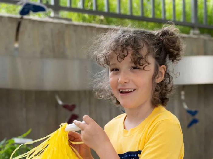 A young girl in a yellow T-shirt with a yellow pompon. She’s outdoors and smiling at the camera. 