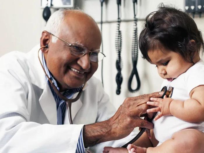 doctor close up listening to baby's heart while baby touches stethoscope while doctor smiles in doctor exam room on exam room table with medical equipment in background
