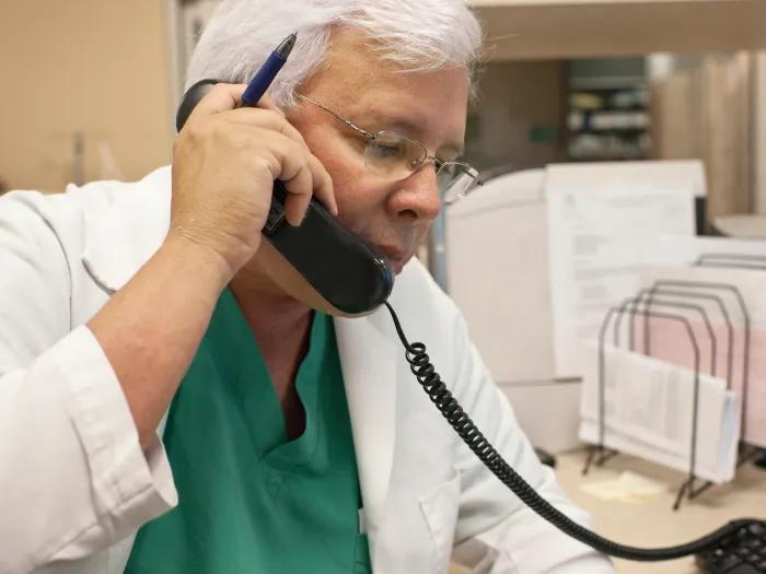 Doctor with white hair and white coat holding a phone to his ear.