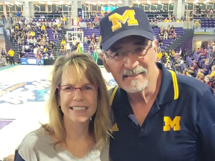 Smiling shot of Julie and Wayne DeLoria wearing U-M gear at a sports event.