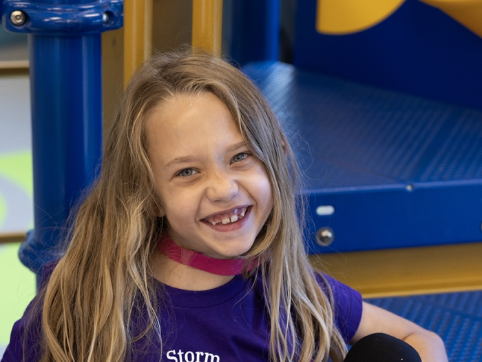 Photo of Evalynn seated on the steps of a piece of play equipment. She has long brown hair and is wearing a purple Calming the Storm of Epilepsy t-shirt.