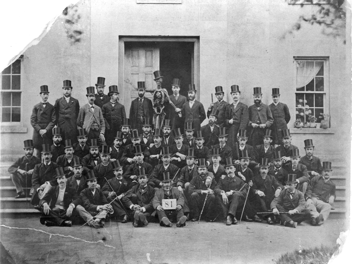 Group of Medical Students from 1881 wearing dark suits and top hats. In back, a student holds up a skeleton wearing a top hat.