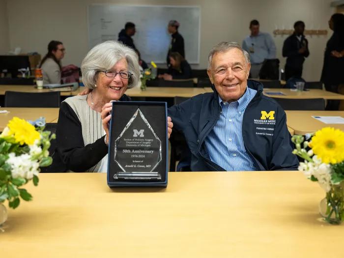 Susan and Arnold Coran are smiling and appear to have just received a plaque that Susan is holding toward the viewer. They are seated behind a table decorated with two vases of flowers.