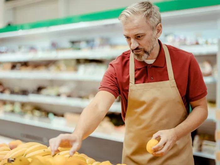 man at grocery store packing produce shelves red shirt beige apron