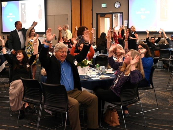A group of about 20 adults seated around several round tables in a large conference room are smiling and raising their arms in what appears to be a celebratory manner.