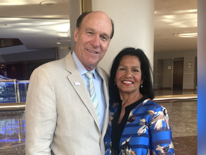 Portrait of Chris and Cindy Harner smiling and turned toward each other. Chris is wearing a light tan suit and Cindy is wearing a blue, black, and tan blouse. They appear to be in an open lobby area.