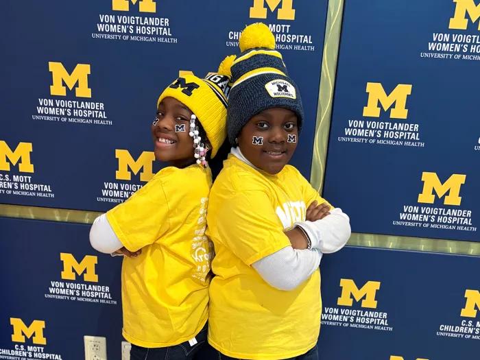 Moriah and Aria stand back-to-back with their arms crossed. They're wearing U-M gear and standing in front of a Mott background.