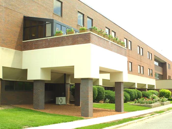 Ground level view of the entrance of Sparrow Hospice House. It is a 3-story brick and concrete building with an extension over the entrance. There are leavy bushes and flowers in front of the building.