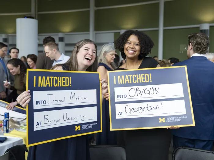 Two U-M medical school students hold up cards announcing their residency matches. The left card reads "Internal Medicine at Boston University." The right card reads "OB/GYN at Georgetown!"