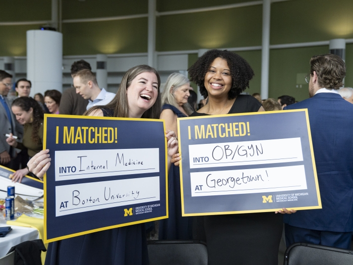Two U-M medical school students hold up cards announcing their residency matches. The left card reads "Internal Medicine at Boston University." The right card reads "OB/GYN at Georgetown!"