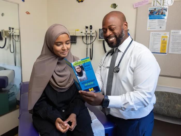 A young, Bengali/Asian, hijabi, Muslim woman looks at a clipboard held by a male, African-American provider. She wears a brown hijab and he wears a grey tie and a stethoscope.