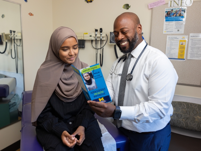 A young, Bengali/Asian, hijabi, Muslim woman looks at a clipboard held by a male, African-American provider. She wears a brown hijab and he wears a grey tie and a stethoscope.