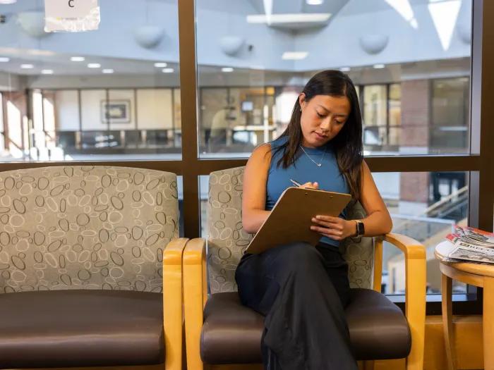 A photo of a young Asian woman sitting in a sunny health care waiting room. She wears a blue sleeveless top and is writing on a clipboard. 