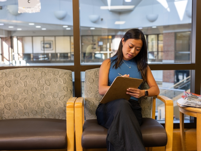A photo of a young Asian woman sitting in a sunny health care waiting room. She wears a blue sleeveless top and is writing on a clipboard. 