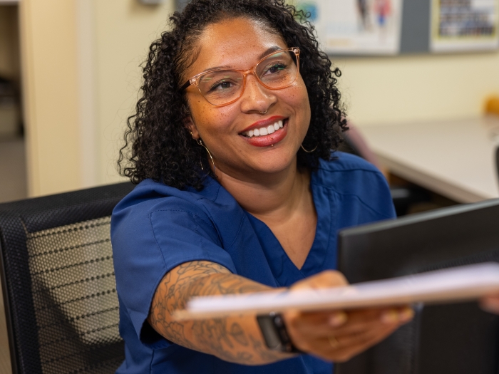 Photo of a tattooed, able-bodied, multiracial woman with a clipboard in a health care office.