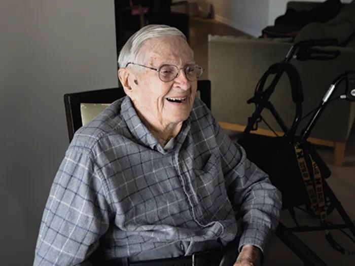 A smiling older man wearing glasses and a plaid shirt is sitting down. There's a walker behind him.