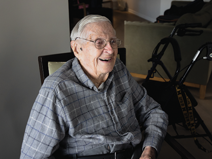 A smiling older man wearing glasses and a plaid shirt is sitting down. There's a walker behind him.