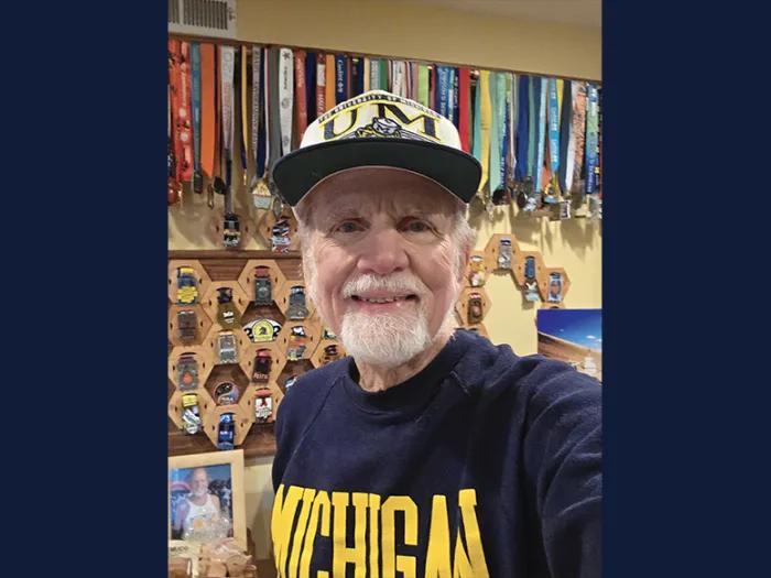 A man wearing a University of Michigan cap and T-shirt poses in front of a wall of marathon medals.
