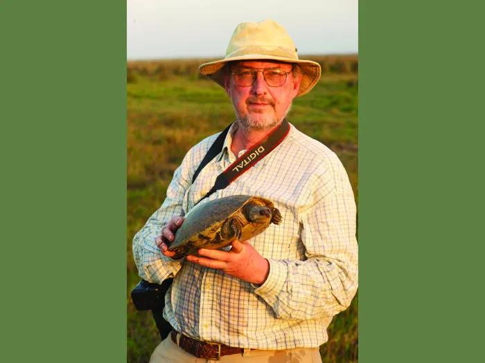 A man in a Panama hat stands outside on a grassy plain holding a large turtle. He has a digital camera around his neck.