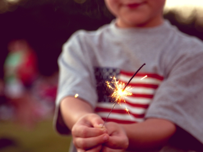 Little boy in American flag t-shirt holds a sparkler near the end of it’s time burning in his hands 