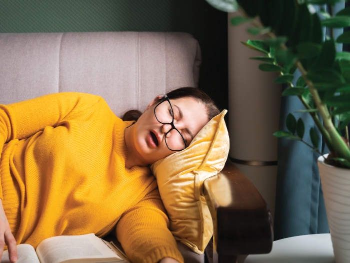 A woman wearing a bright yellow sweater is sleeping on her side on a couch. Her mouth is open and her glasses are askew. Her fingers are resting on an open book.