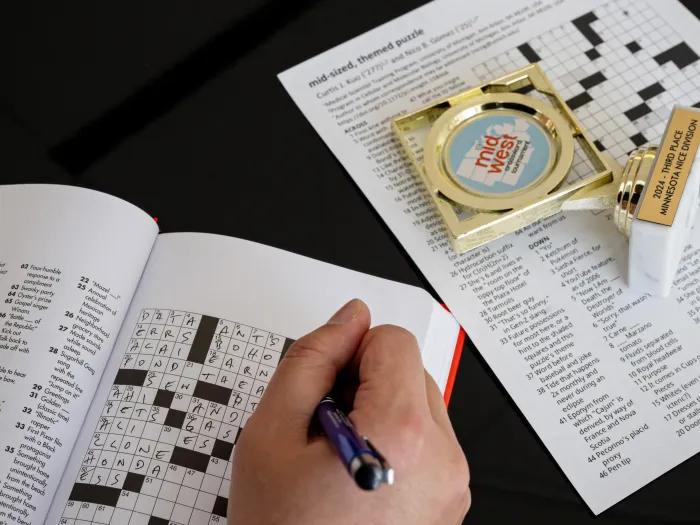 A person filling out a crossword puzzle. Only their hand is visible. There's also a small trophy on the right side that commemorates winning a prize at a crossword puzzle competition.